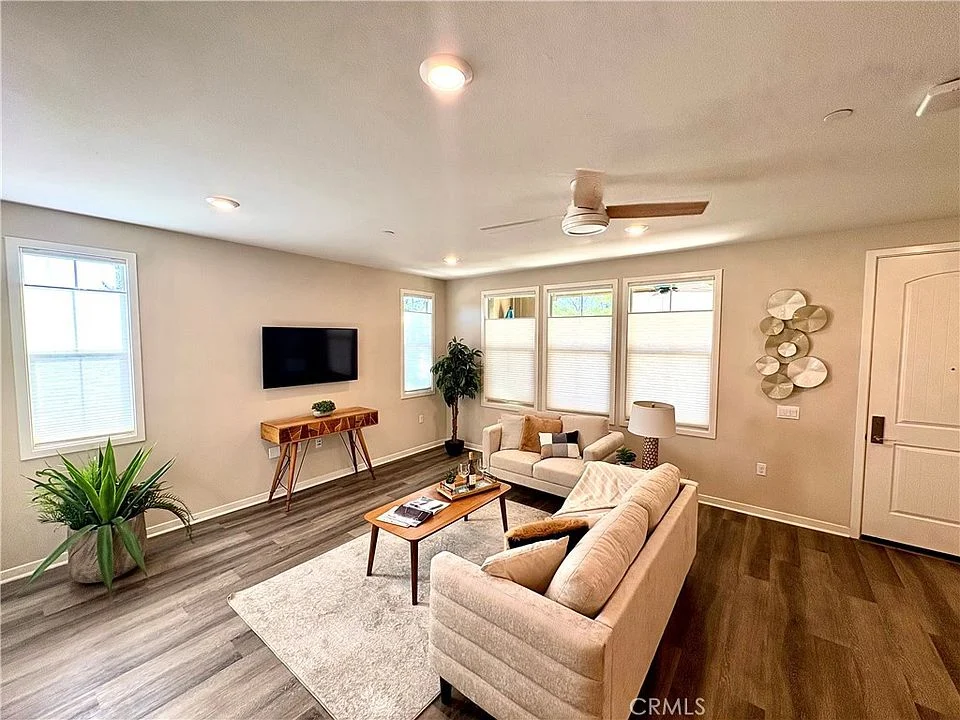 Living room with sofa, wood table, wall TV, plants, and bright windows.