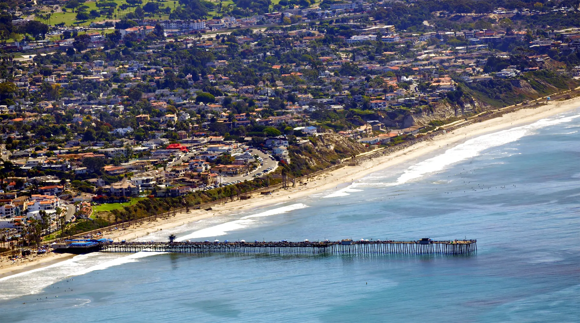 Aerial view of pier in ocean with beach and nearby homes, real estate.