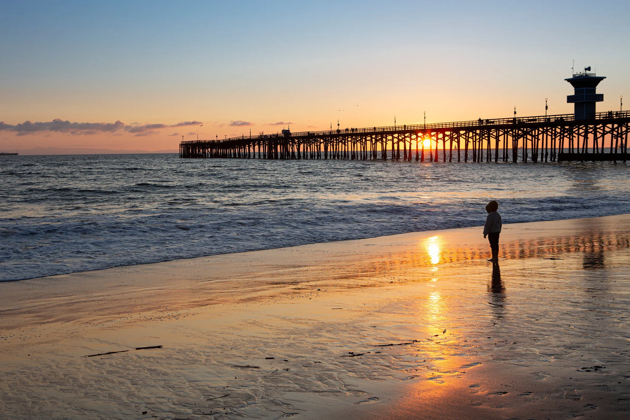 Child standing on beach with pier and ocean waves in background.
